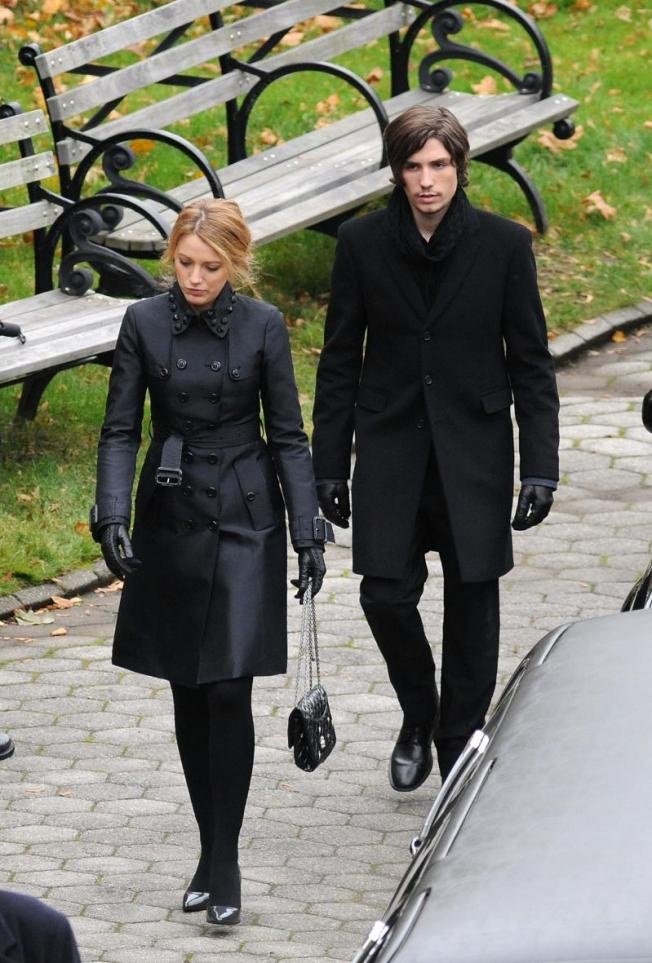 Serena van der Woodsen walking through a park in a belted black trench coat beside Aaron Rose